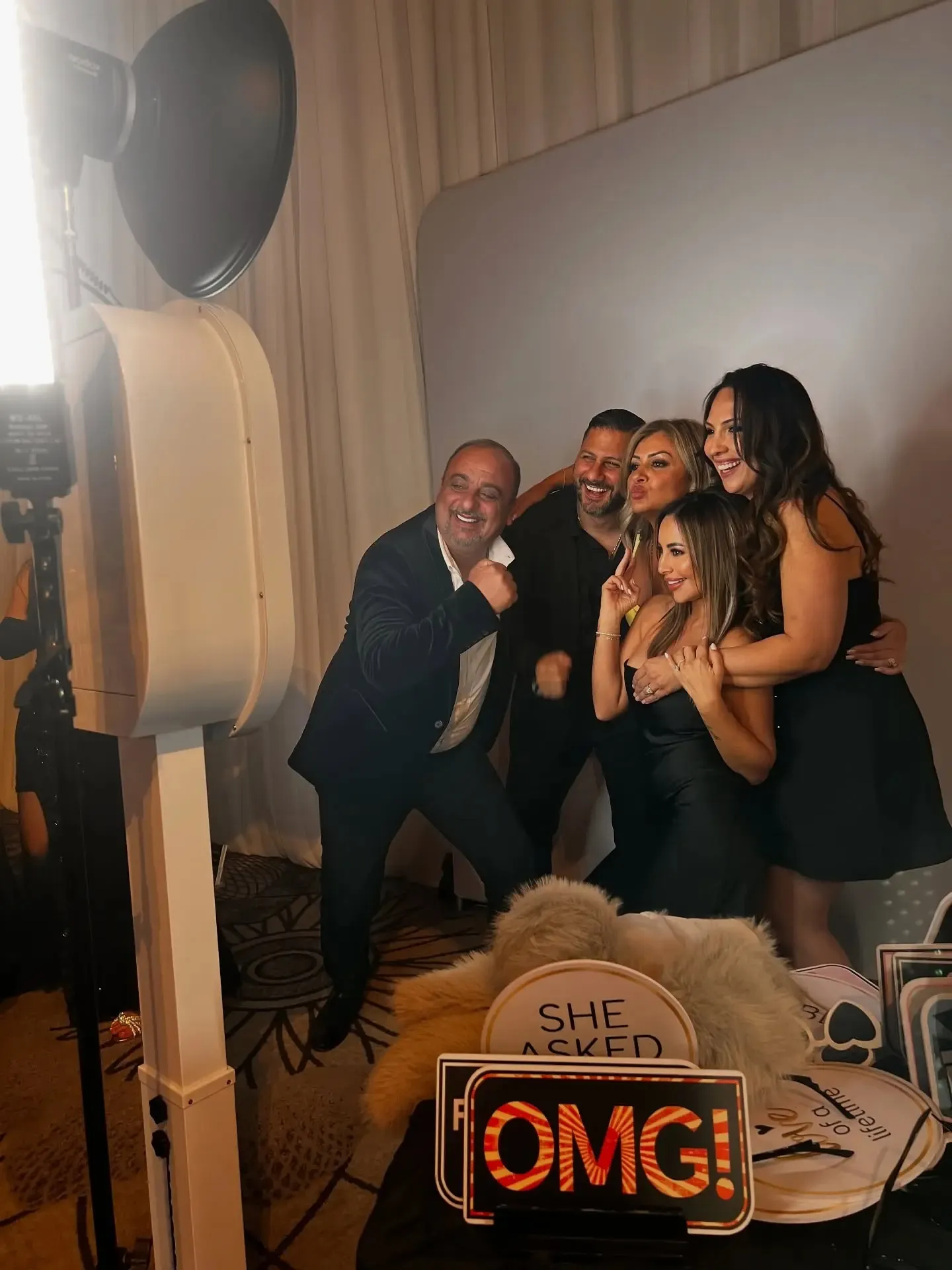 Group of four people posing joyfully in front of a 360 video booth, with a bright light and a sign reading "SHE ASKED OMG!" in a festive event setting.