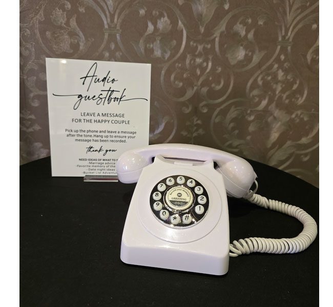 Audio guestbook setup featuring a vintage white rotary phone and a sign instructing guests to leave messages for the happy couple, set against an elegant backdrop.