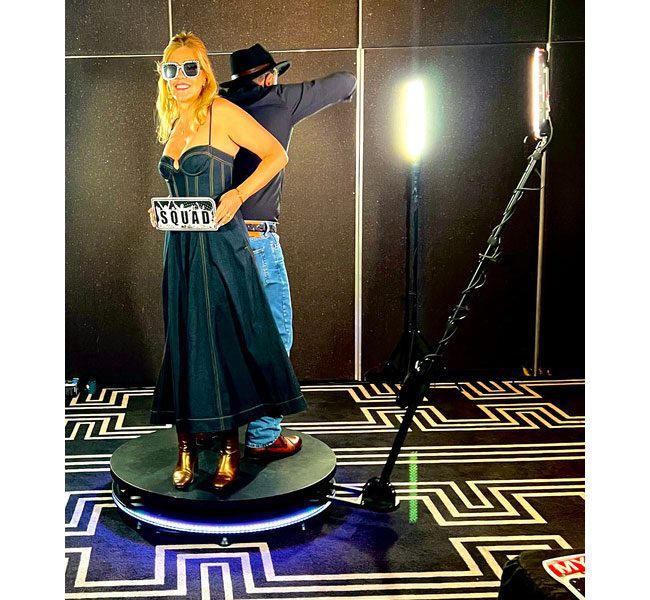 Woman in a black dress with sunglasses holding a "SQUAD" sign, standing on a rotating platform with a photographer behind her, illuminated by studio lights, showcasing Halo 360 video booth experience.