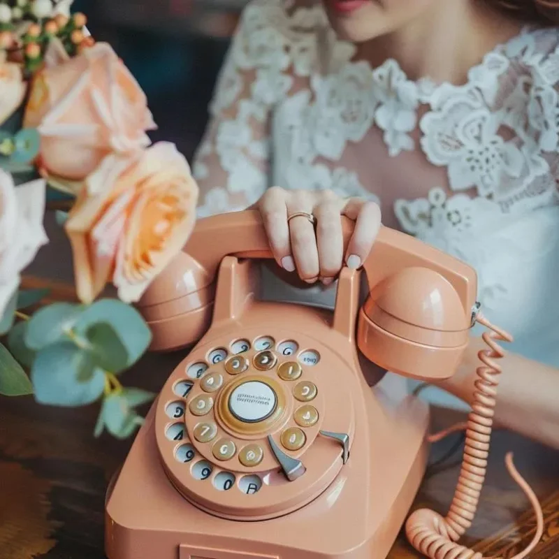 Woman in lace dress holding a vintage pink rotary phone with floral arrangement nearby, symbolizing nostalgia and personal connection.