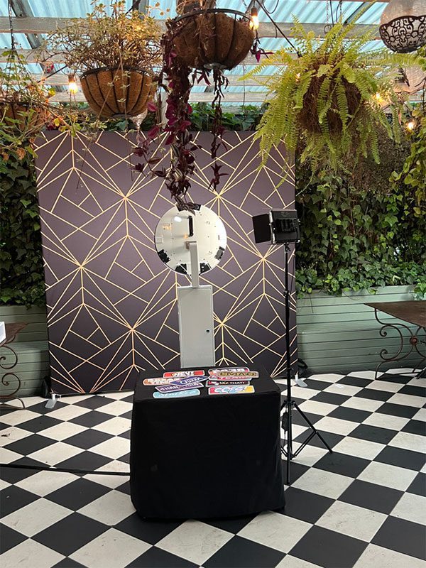 360 photo booth setup with black and gold geometric backdrop, surrounded by greenery and hanging plants, featuring a table with colourful props, ideal for capturing memorable moments at events.