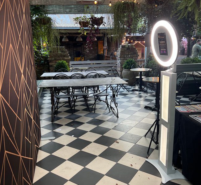 Halo 360 photo booth setup featuring a circular light stand and tables in a stylish outdoor venue with a checkered floor, surrounded by greenery and decorative elements.