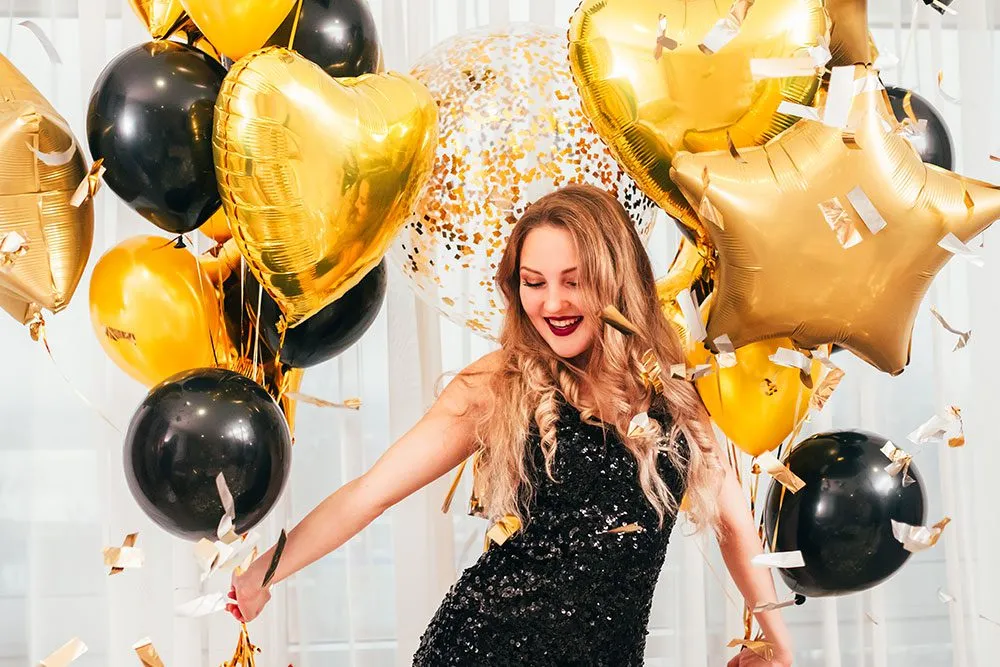 Woman in a black sequined dress celebrating with gold and black balloons, surrounded by confetti, embodying a festive atmosphere for events like video guest book services.
