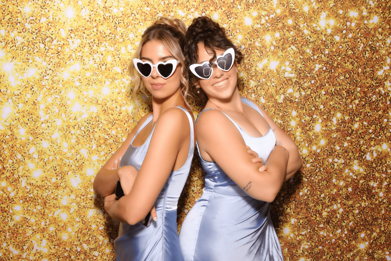 Two women in elegant blue dresses and heart-shaped sunglasses posing back-to-back against a sparkling gold backdrop, capturing a fun moment at a wedding photo booth experience.