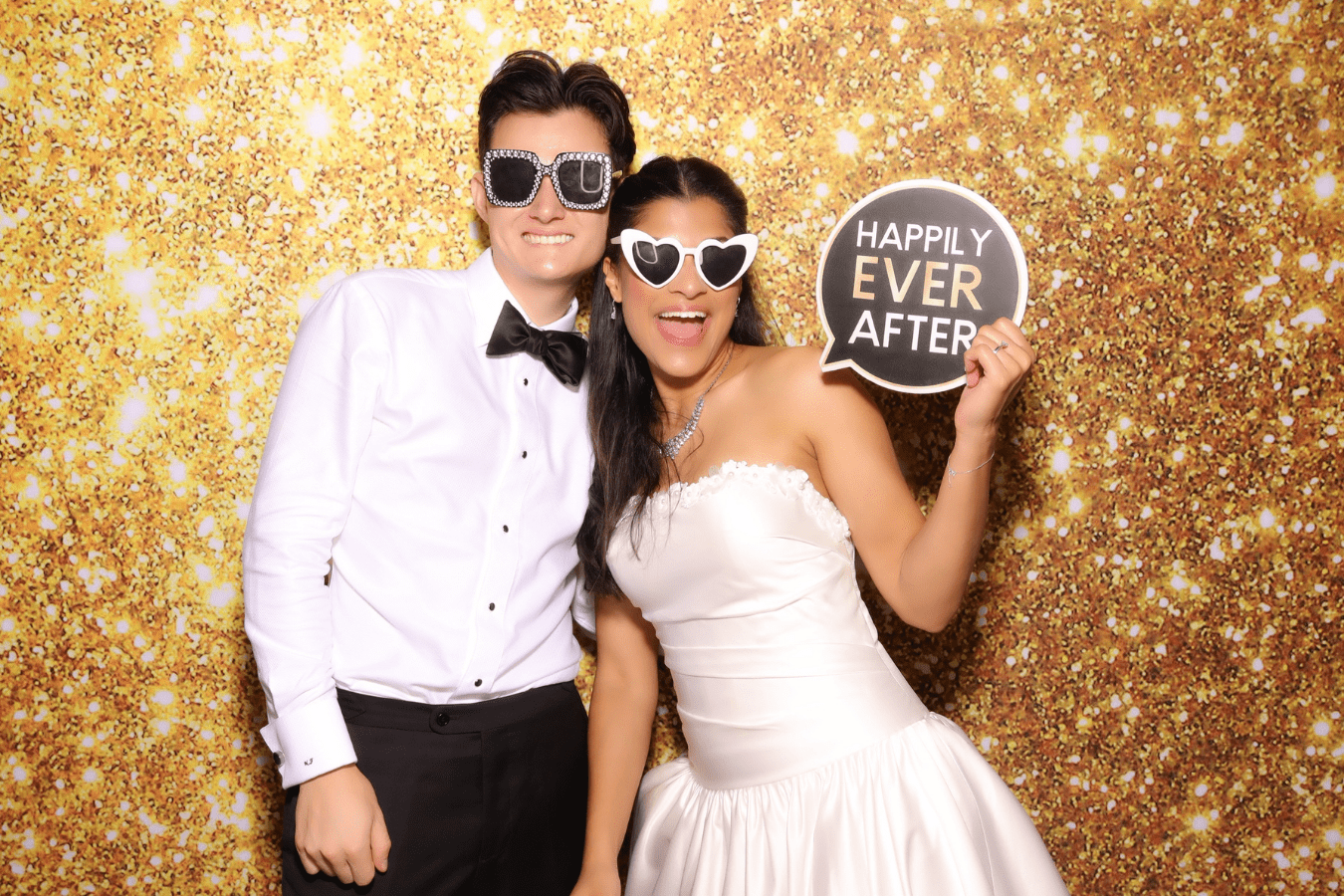 Couple posing with playful sunglasses and a "HAPPILY EVER AFTER" sign, set against a sparkling gold backdrop, capturing a joyful moment at a wedding photo booth experience.