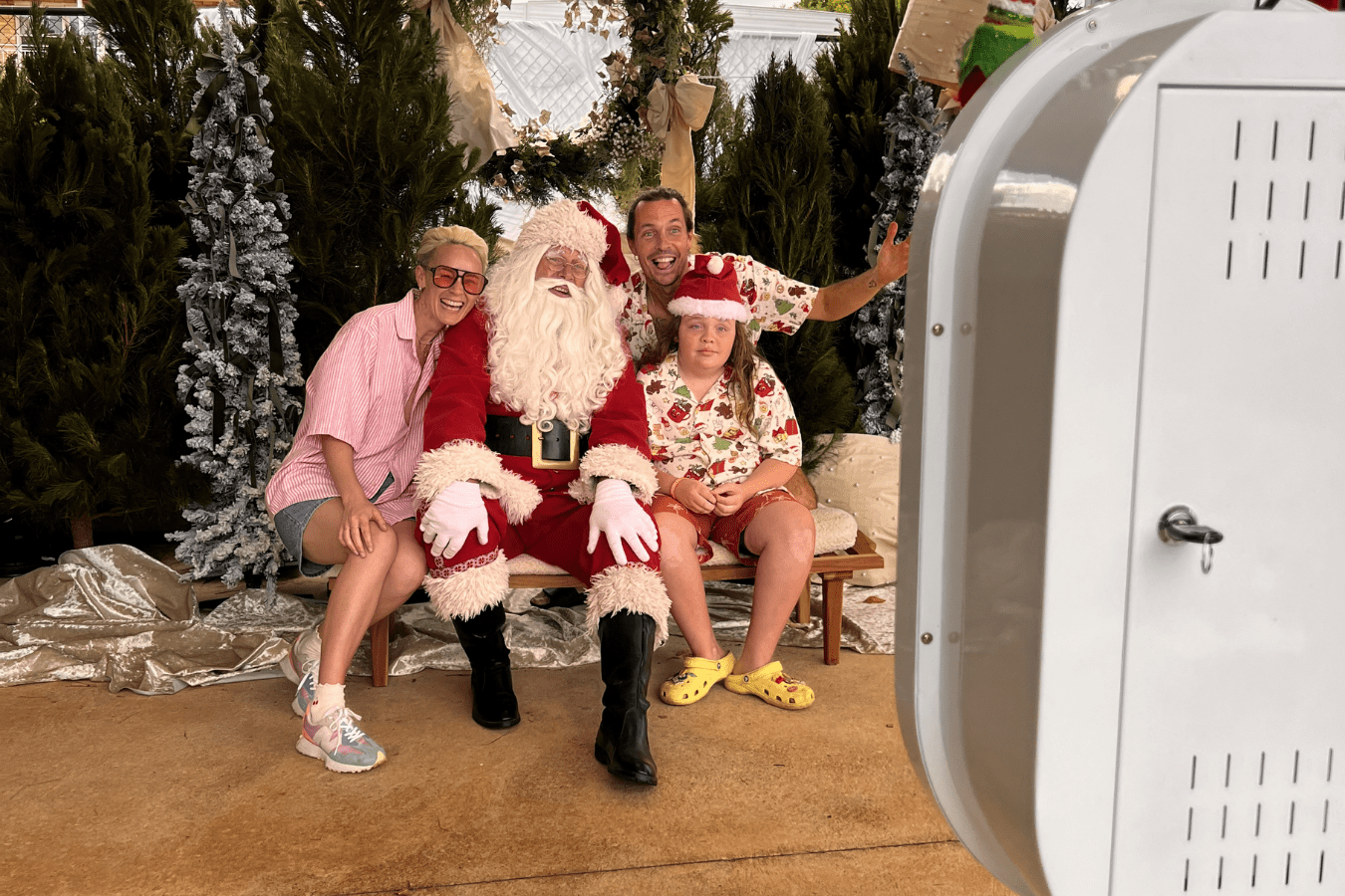 Family posing with Santa Claus at a festive photo booth setup, featuring Christmas decorations and a digital photo booth in the foreground.