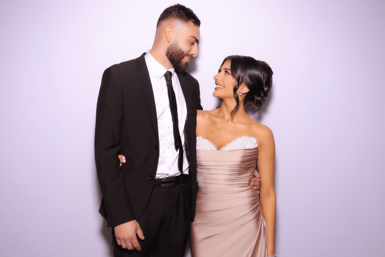 Couple smiling and posing together in formal attire, suitable for a Quinceañera celebration, with a soft purple background.