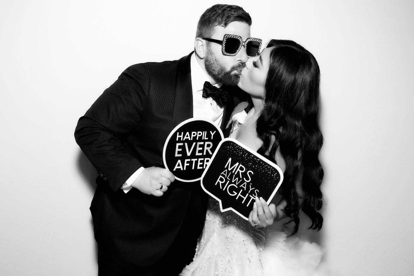 Couple kissing while holding fun signs "HAPPILY EVER AFTER" and "MRS. ALWAYS RIGHT," showcasing a joyful moment at a wedding photo booth.
