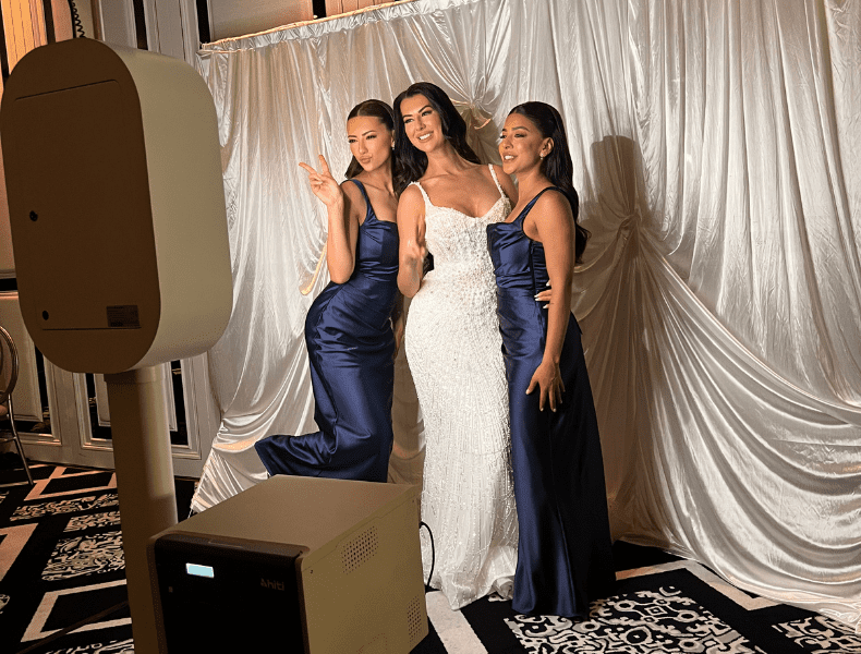 Three women posing joyfully in elegant dresses in front of a photo booth at a wedding, showcasing a premium photo experience with a sleek setup and polished backdrop.