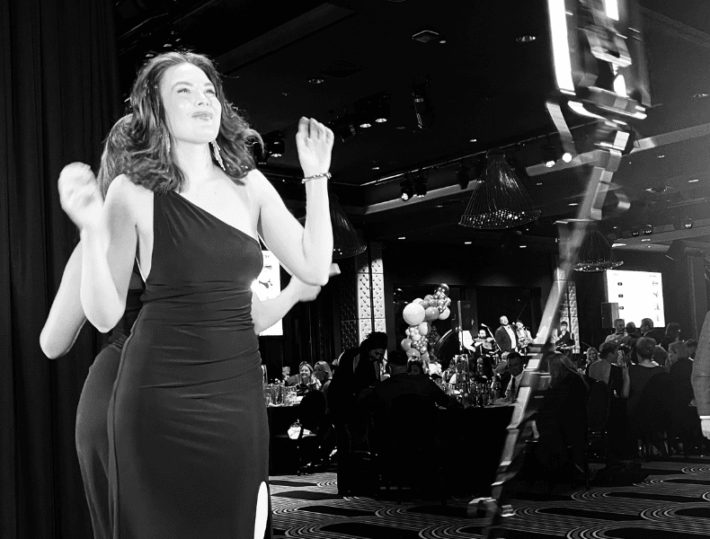 Woman in elegant black dress posing joyfully at an event, showcasing a vibrant atmosphere with guests and decor in the background, emphasizing premium wedding photo booth experiences.