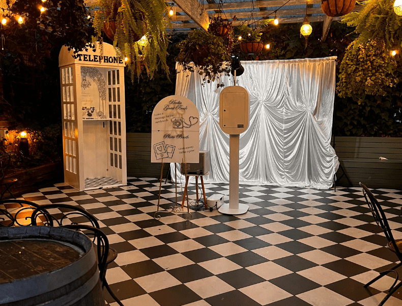 Photo booth setup featuring a vintage telephone booth, a white backdrop with drapery, and a sleek photo booth station on a checkered floor, surrounded by greenery and warm ambient lighting, ideal for wedding celebrations.