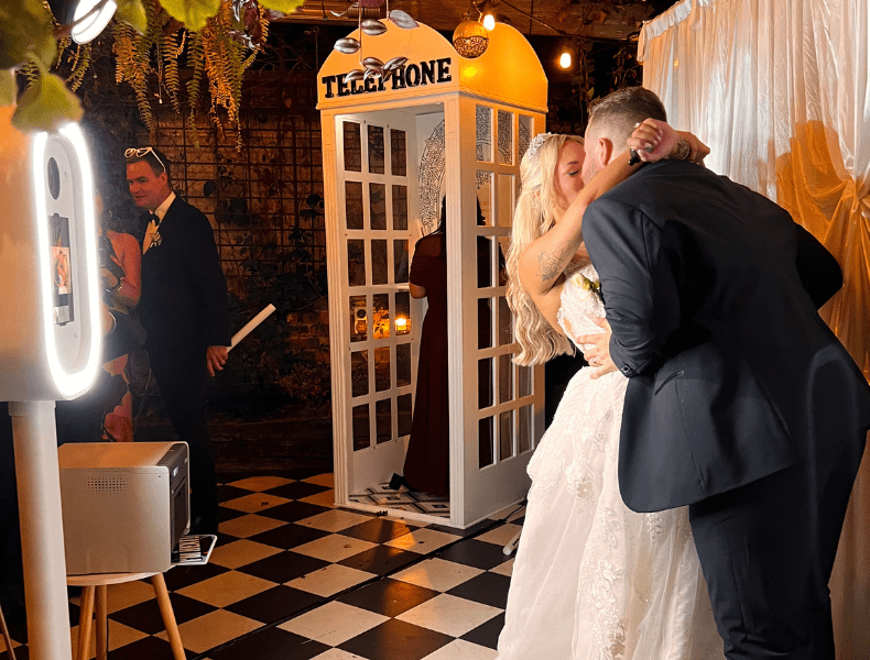 Bride and groom kissing in front of a vintage telephone booth photo booth, with guests nearby, showcasing a stylish wedding setup with professional lighting and decor.